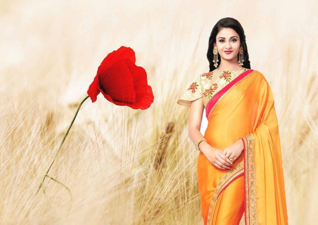 Woman in a vibrant orange saree with floral blouse, standing in a wheat field with a red flower, highlighting the saree collection for every occasion